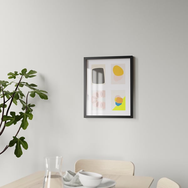 Framed abstract art hangs on a gray wall above a wooden table with a glass, plates, and a plant.