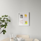 Modern art prints in floating frames above wooden table with chair, plant, glass, and bowl.
