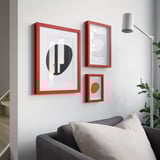 Three red RÖDALM frames on white wall with abstract art. One above grey couch with white pillows. White floor lamp and green plant nearby.