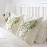 Two decorative pillows with green butterfly designs sit on a white bed. A small vase with white flowers is on the bedside table.