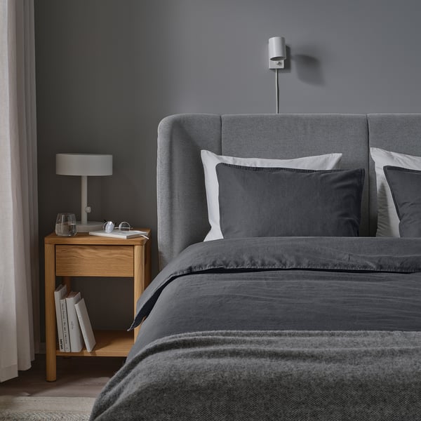 Minimalist bedroom with grey duvet, pillows, wooden nightstand, wall light, small lamp, books. Dark grey walls, light curtains.