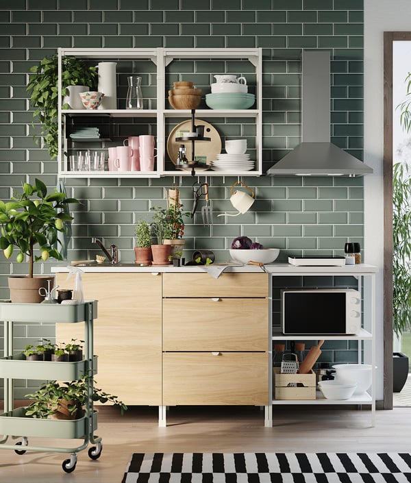 Modern kitchen with light wood cabinets, green tiles, stainless steel hood, plants, and open shelving with dishes and cups.