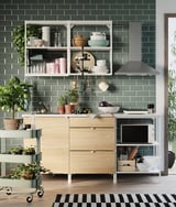 Modern kitchen with light wood cabinets, green tiles, stainless steel hood, plants, and open shelving with dishes and cups.
