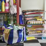 Colorful bags filled with cleaning supplies and gloves, beside a tall stack of assorted books on a cluttered desk.