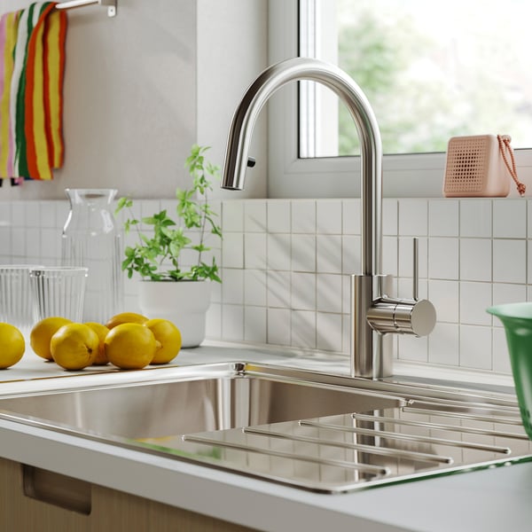 Modern kitchen sink with stainless steel SALLSJÖN tap, curved design, surrounded by lemons, herbs, and colorful towels.