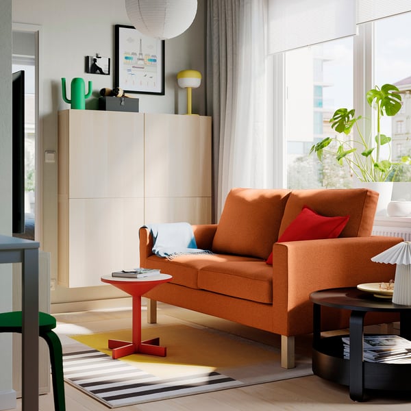 Orange SALTSJÖBADEN sofa, large windows, wooden floor, colourful rug, and plants, with modern decor and green cabinet.