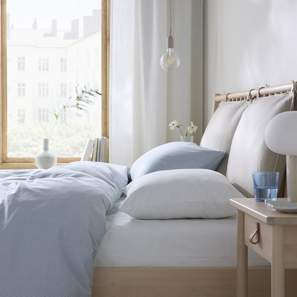 Bright, minimalist bedroom with light blue bedding, wooden nightstand, hanging bulb, and city view.