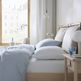 Bright, minimalist bedroom with light blue bedding, wooden nightstand, hanging bulb, and city view.