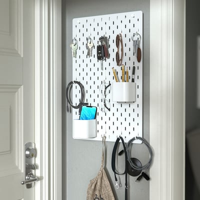 A white SKÅDIS pegboard organizer holds multiple items such as keys, pens, headphones, and a phone, offering a neat and accessible storage solution.