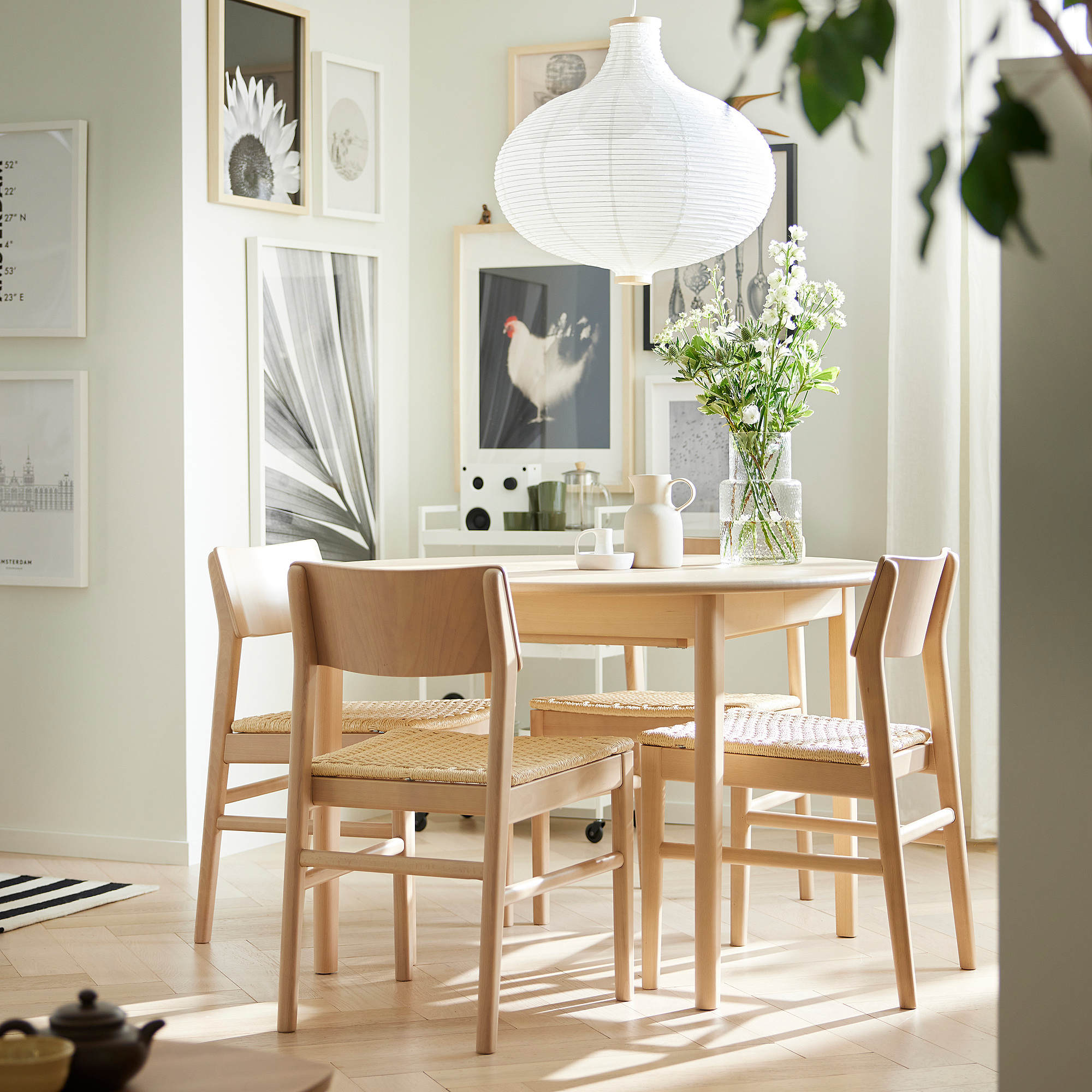 Dining room with wooden table, chairs, and white flowers. Central table under round lantern, framed art on walls.