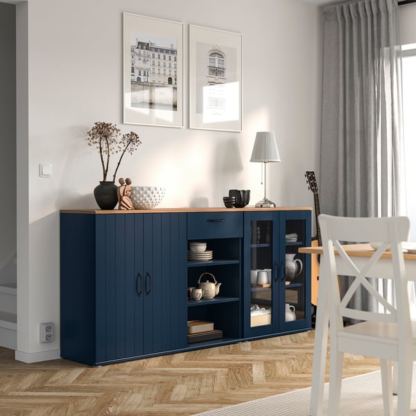 Modern dining room with blue SKRUVBY sideboard, glass doors, and china. Gray curtains, white prints, wooden floor.