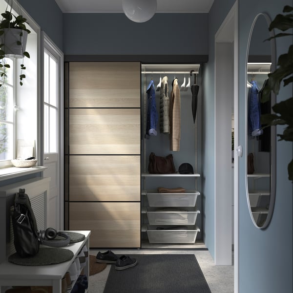 Small walk-in closet with light wood sliding doors, hanging clothes, white shelves, and gray mesh baskets. Round mirror and plants beside the door.