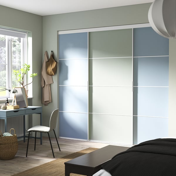 Modern bedroom with large, light blue, sliding wardrobe. It a minimalist desk, chair, and decorative plants.