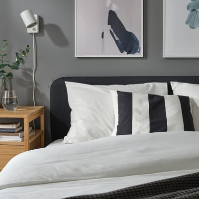 Modern bedroom with black & white striped bed pillows, grey wall art, and a wooden bedside table with books and greenery.