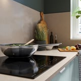 Modern kitchen with induction cooktop. Black wok pan, wooden utensils, and fresh vegetables. Light-colored counters.