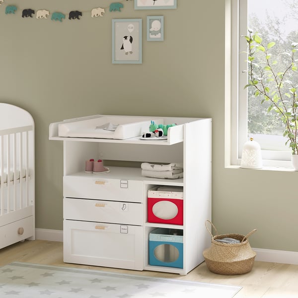 In a nursery, a white nursery table with shelves and drawers holds baby items. Decorated with toys and books, it stands near a window with plants.