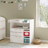 In a nursery, a white nursery table with shelves and drawers holds baby items. Decorated with toys and books, it stands near a window with plants.