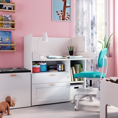 Kids study area with white SMÅSTAD desk, blue chair, books, and toys. Pink walls with windows and curtains.