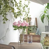 Vase of pink roses on table. Surrounded by plants, outdoor setting.