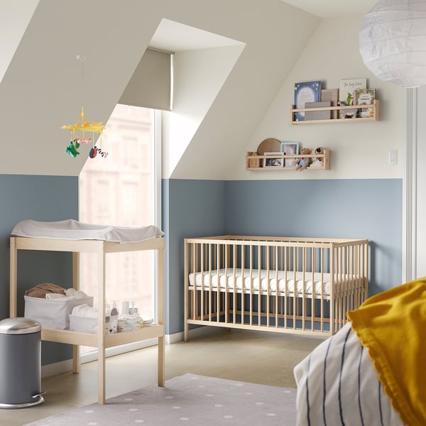 Baby nursery with light blue walls, light wood crib, changing table, shelves, and yellow accents.