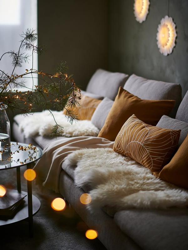 Cozy living room with grey SÖDERHAMN sofa, brown pillows, sheepskin, and christmas decorations - including hanging lights and pine branches.