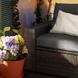 Modern outdoor chair with grey cushions and white pillow, next to potted purple and white flowers, set against a yellow wall and blue shutters.