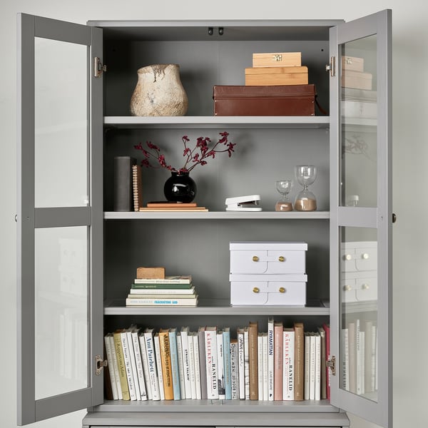 A modern gray glass-door cabinet displaying books, decorative vases, and storage boxes on three shelves.