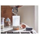 A person shakes powder into a drink or batter using a gradient-marked shaker on a kitchen counter.