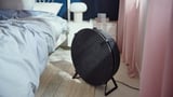 Black ikea STARKVIND air purifier beside a bed with gray bedding and a white lamp on a nightstand.