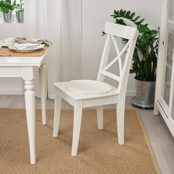 White sheepskin chair pad on white wooden chair near dining table with plate, cutlery, beige rug, and plant.