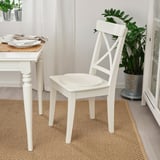 White sheepskin chair pad on white wooden chair near dining table with plate, cutlery, beige rug, and plant.