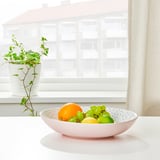 White bowl of fruits on a table by a window with a plant.