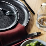 A table setting with black cutlery, plates, and a glass. Fork, knife, and spoon rest on plates; green dish below.