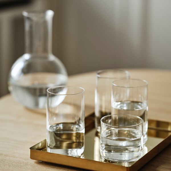 Four sleek, modern glasses on a gold tray, filled with water. Behind them, a large carafe in a minimalist kitchen.