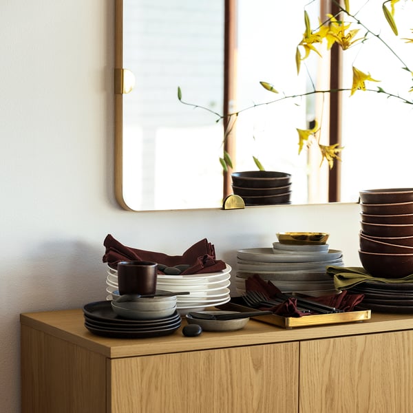 Wooden cabinet with stacked plates, bowls, and cloths in earthy tones against white wall featuring a mirror and a large plant.