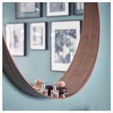 A round walnut-framed mirror with a bottom shelf holds perfume bottles and a ring box against a blue wall adorned with picture frames.