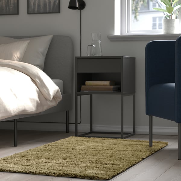 Bedroom with grey bed, blue chair, olive rug, black side table with books and pitcher.