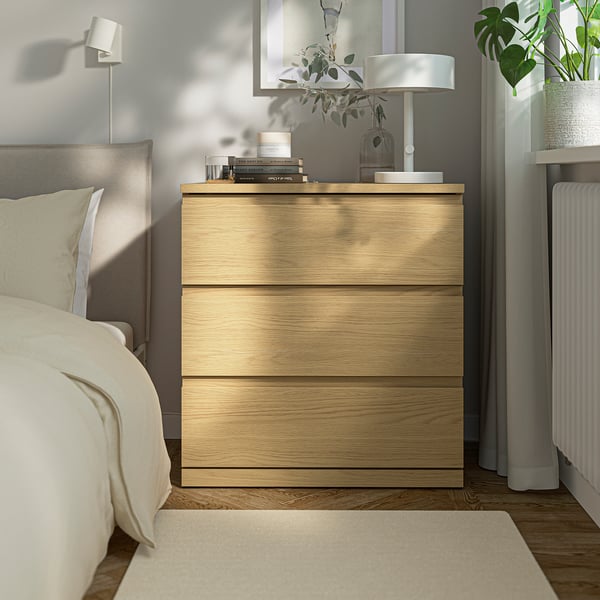 A light wooden bedside chest, smaller than the bed, with a lamp, plants, and books on top.