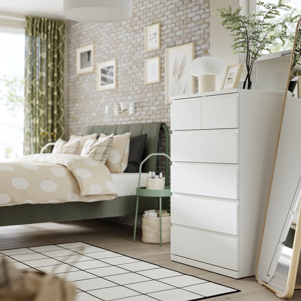 Modern bedroom with storklintha white dresser, bed, mirror, plants, lamp, and decor.