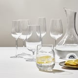 Six STORSINT crystal glasses of various sizes, one filled with lemonade, surrounded by lemon slices, and a carafe on a white tablecloth.