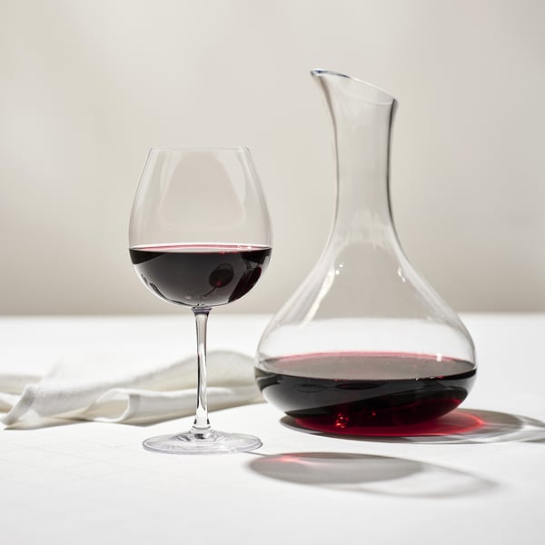 A clear wine glass and decanter on a table. The glass is half full of red wine, and the decanter holds more wine, enhancing its flavor and aroma.