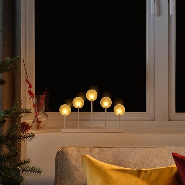 Cozy indoor scene STRÅLA candle holder with warm led lights on a windowsill, flanked by festive decorations and a yellow pillow.