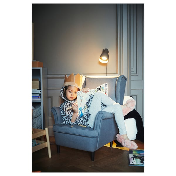 A child sits in a gray STRANDMON armchair, wearing a crown and pink slippers, holding a stuffed toy.