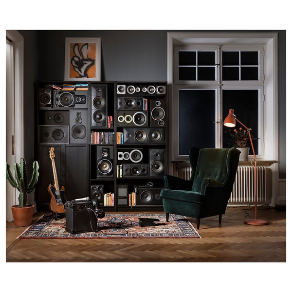 Home music room with large speaker shelves, green chair, guitar, vintage amp, vintage rug, cactus, lamp on wooden floor.