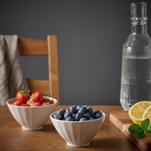Two white ceramic bowls filled with strawberries and blueberries on wooden table, with bottle and lemon slice nearby.
