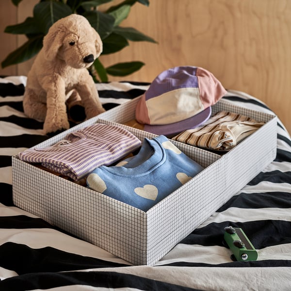 A grid-patterned storage box holds folded baby clothes and a hat on a striped bed. A stuffed animal and a green toy lie nearby.