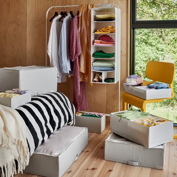 Bedroom with wooden walls, striped bedding, and white storage boxes. Clothes hang from a rack, and a yellow chair sits near a window.