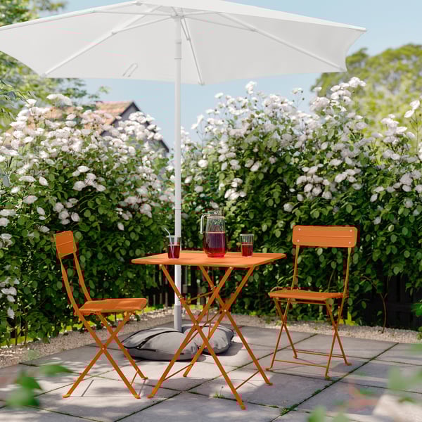 A serene garden setting with a white umbrella, orange foldable table, and chairs surrounded by blooming bushes.