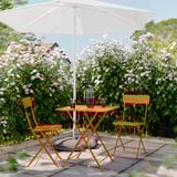 A serene garden setting with a white umbrella, orange foldable table, and chairs surrounded by blooming bushes.