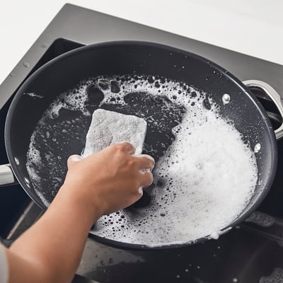 Person scrubbing a pan with a dual-sided sponge; one side soft, one side coarse, suitable for cleaning dishes and tough stains.